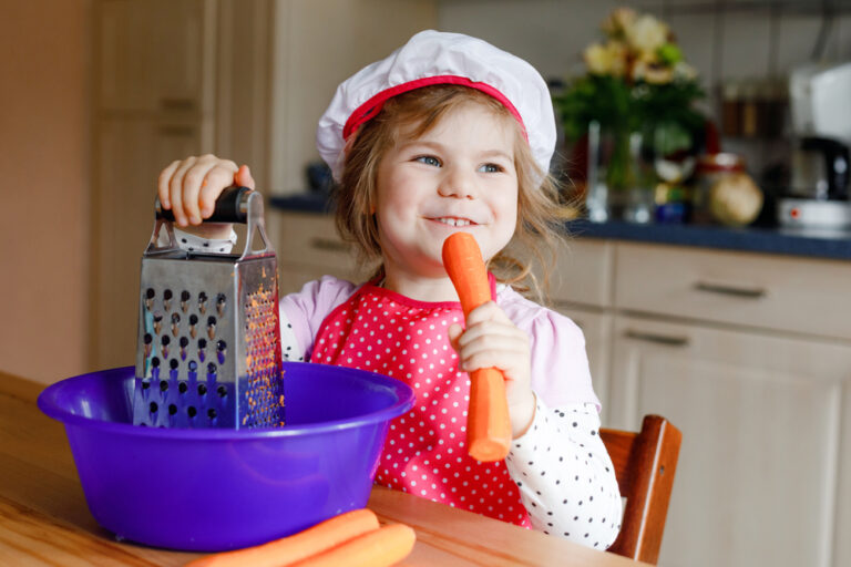 Cute little preschool girl grate carrots for baking carrot cake or cookies. Happy toddler child in apron and chef hat help in the kitchen prepare healthy salad lunch, indoors. Healthy food with kids