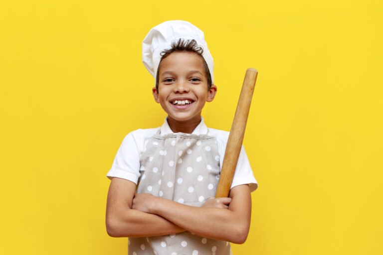 african american boy in chef's uniform and hat holding kitchen items and smiling on yellow isolated background, teenager child in apron with crossed arms looking at camera